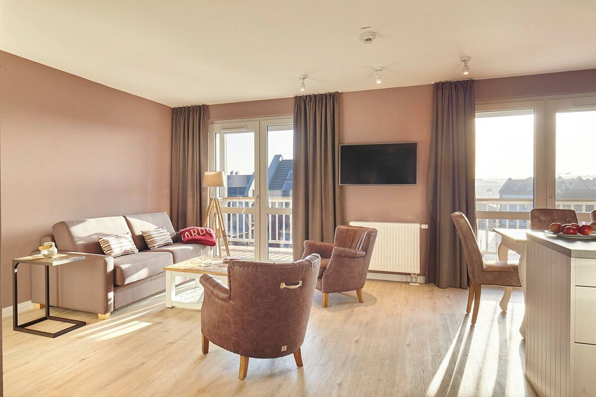 Un salon moderne comprenant un canapé, des fauteuils, une table basse et un coin repas avec des chaises et une table. Un téléviseur à écran plat est fixé au mur, et de grandes fenêtres sont agrémentées de rideaux.