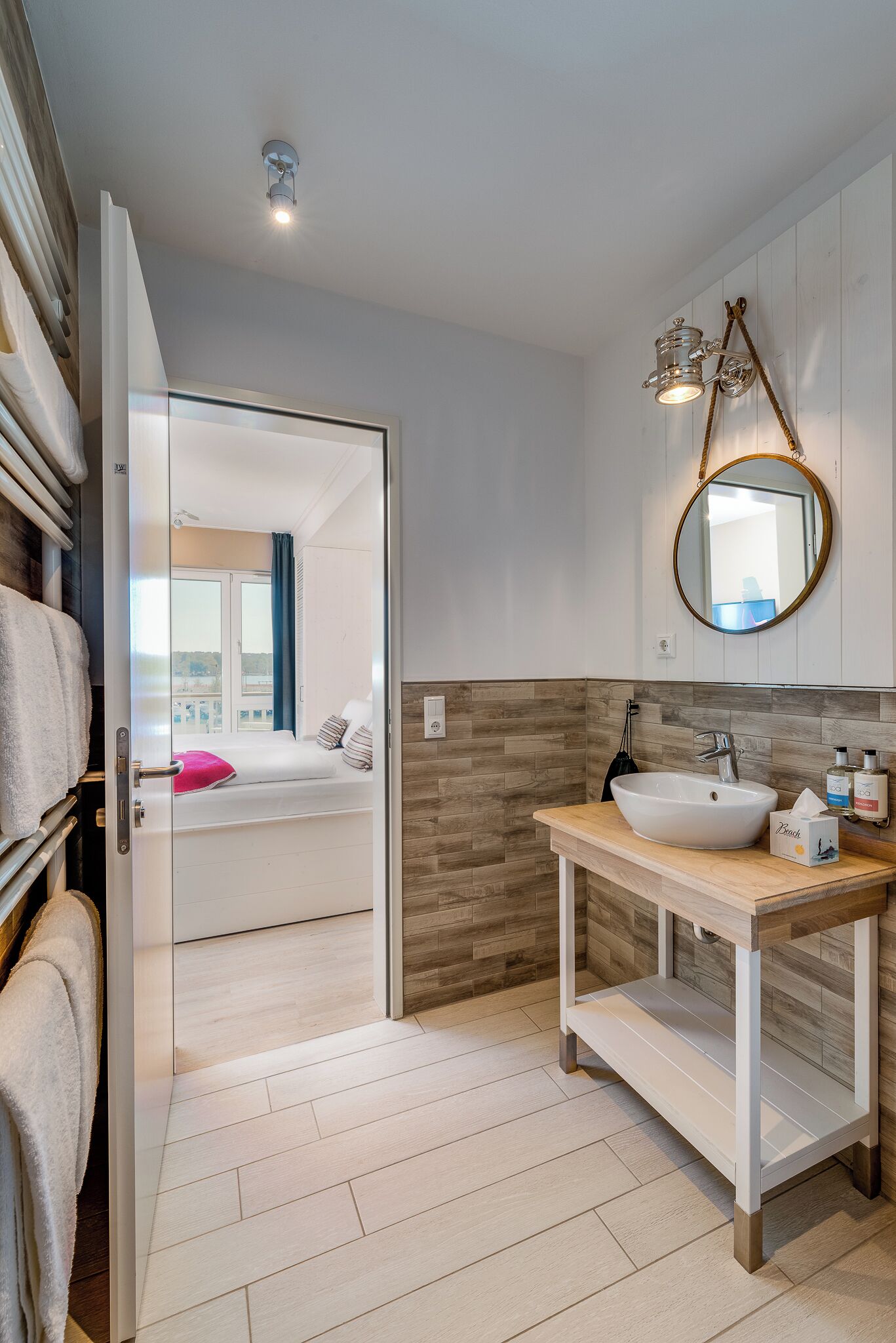 A modern bathroom with a white sink, wooden vanity, and a large mirror. A bed with a pink pillow is visible through an open door.