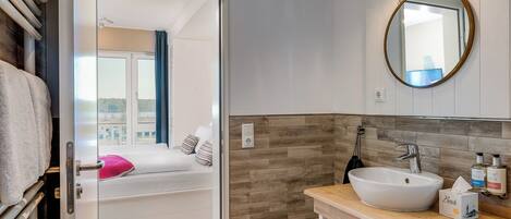 A modern bathroom with a white sink, wooden vanity, and a large mirror. A bed with a pink pillow is visible through an open door.