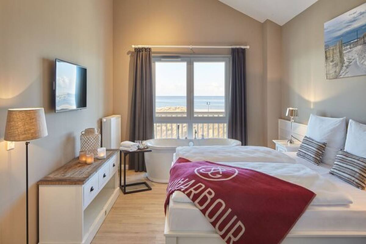 Une chambre d’hôtel avec un lit, une baignoire, une fenêtre avec des rideaux, une table de chevet avec une lampe et une vue sur la mer.