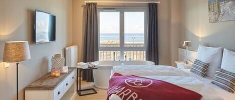 Habitación de hotel con cama, bañera, ventana con cortinas, mesita de noche con lámpara y vista al mar.