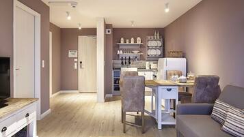A modern living room with a sofa, a dining table with chairs, and a kitchen area with shelves and appliances.