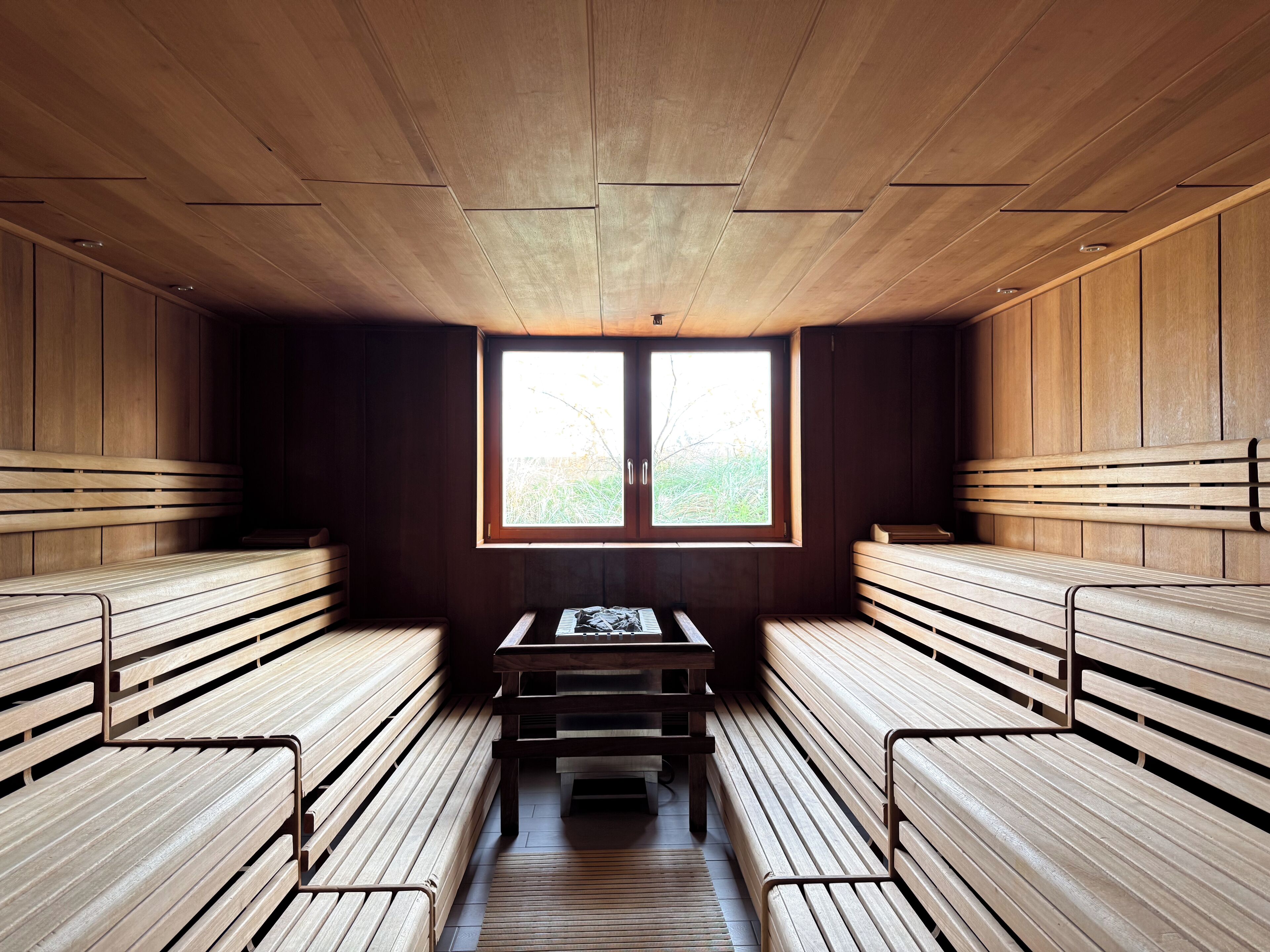 A sauna room with wooden benches and a table, a window with a view of greenery, and a wooden ceiling.