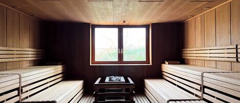 A sauna room with wooden benches and a table, a window with a view of greenery, and a wooden ceiling.