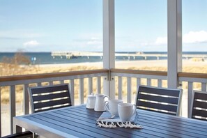 Una mesa con dos sillas y un juego de tazas y jarra blancos en una terraza de madera con vista a una playa y un muelle.