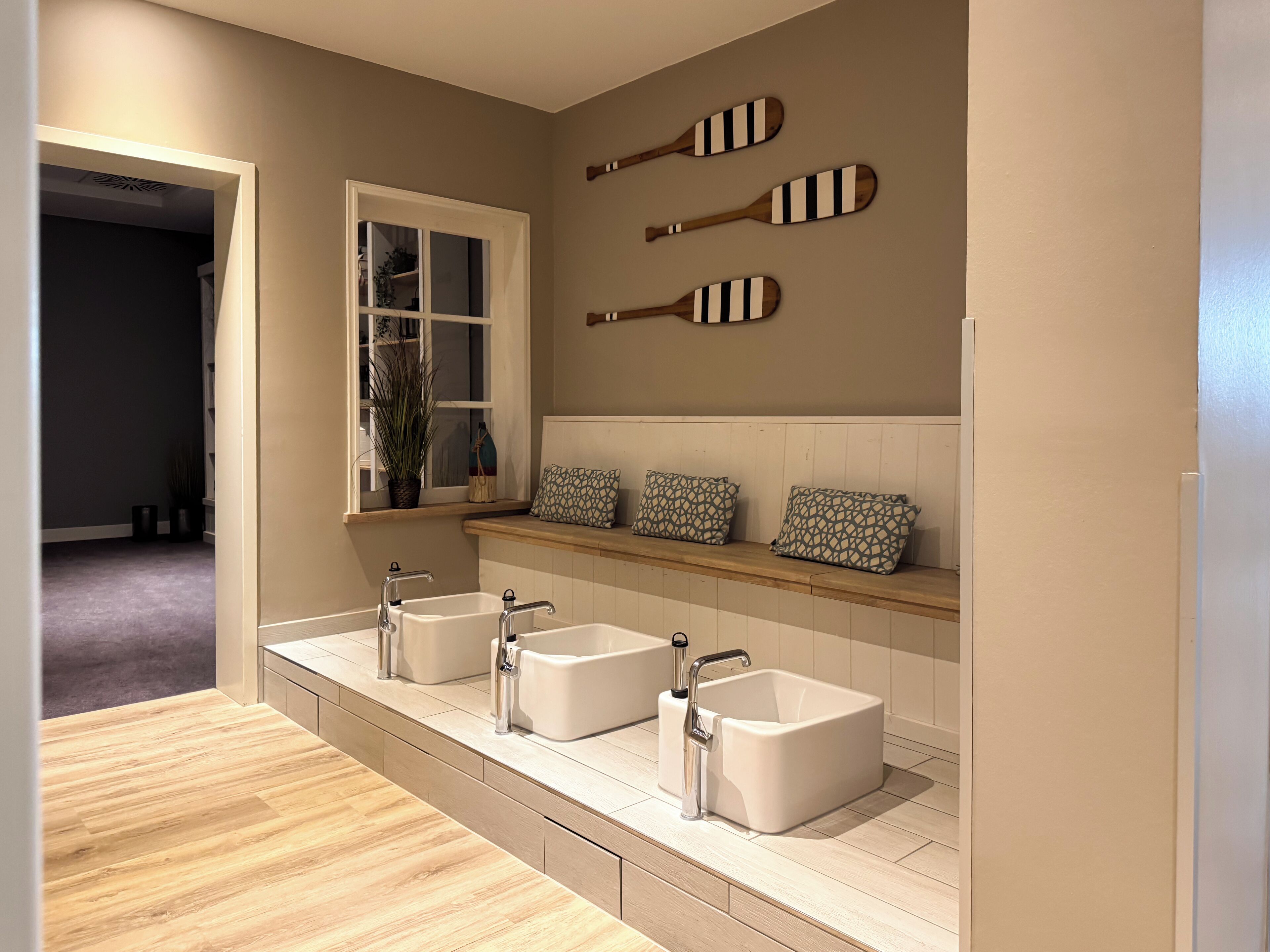 Een badkamer met drie witte wasbakken, een raam en drie houten paddles aan de muur.