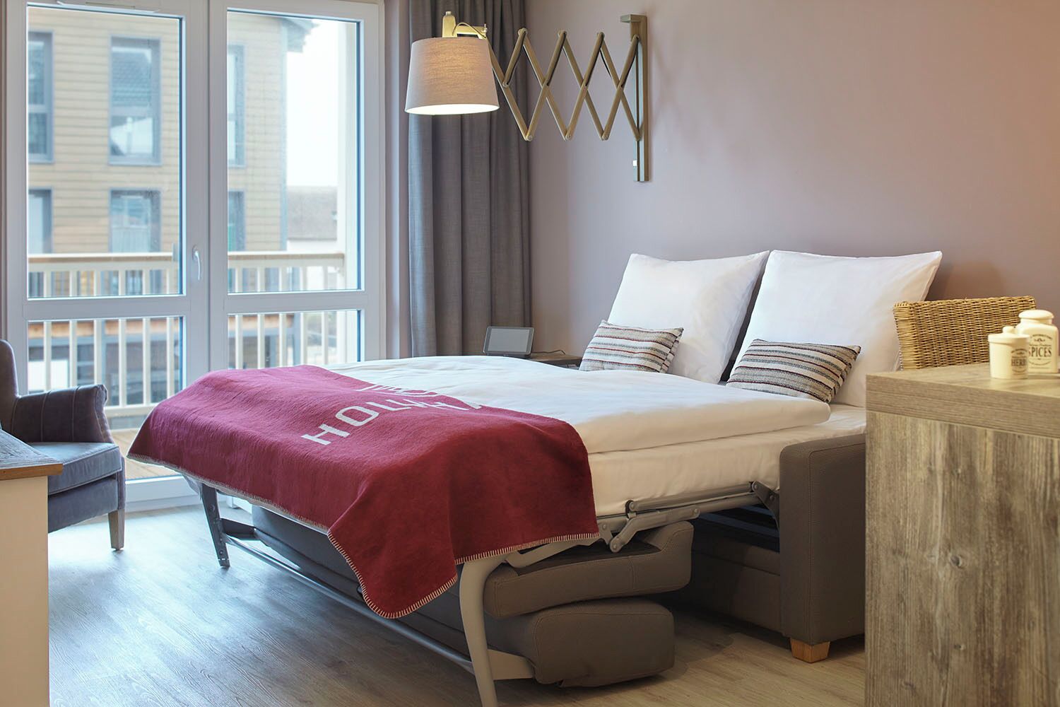 Une chambre d’hôtel avec un lit, une table de chevet, une lampe et une vue sur les bâtiments, visible à travers de grandes fenêtres.