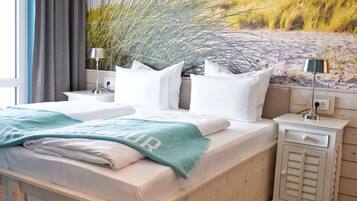 Een net opgemaakt bed met witte lakens en een blauwgroene plaids, een nachtkastje met een lamp, en een wandplank met decoratieve planten.