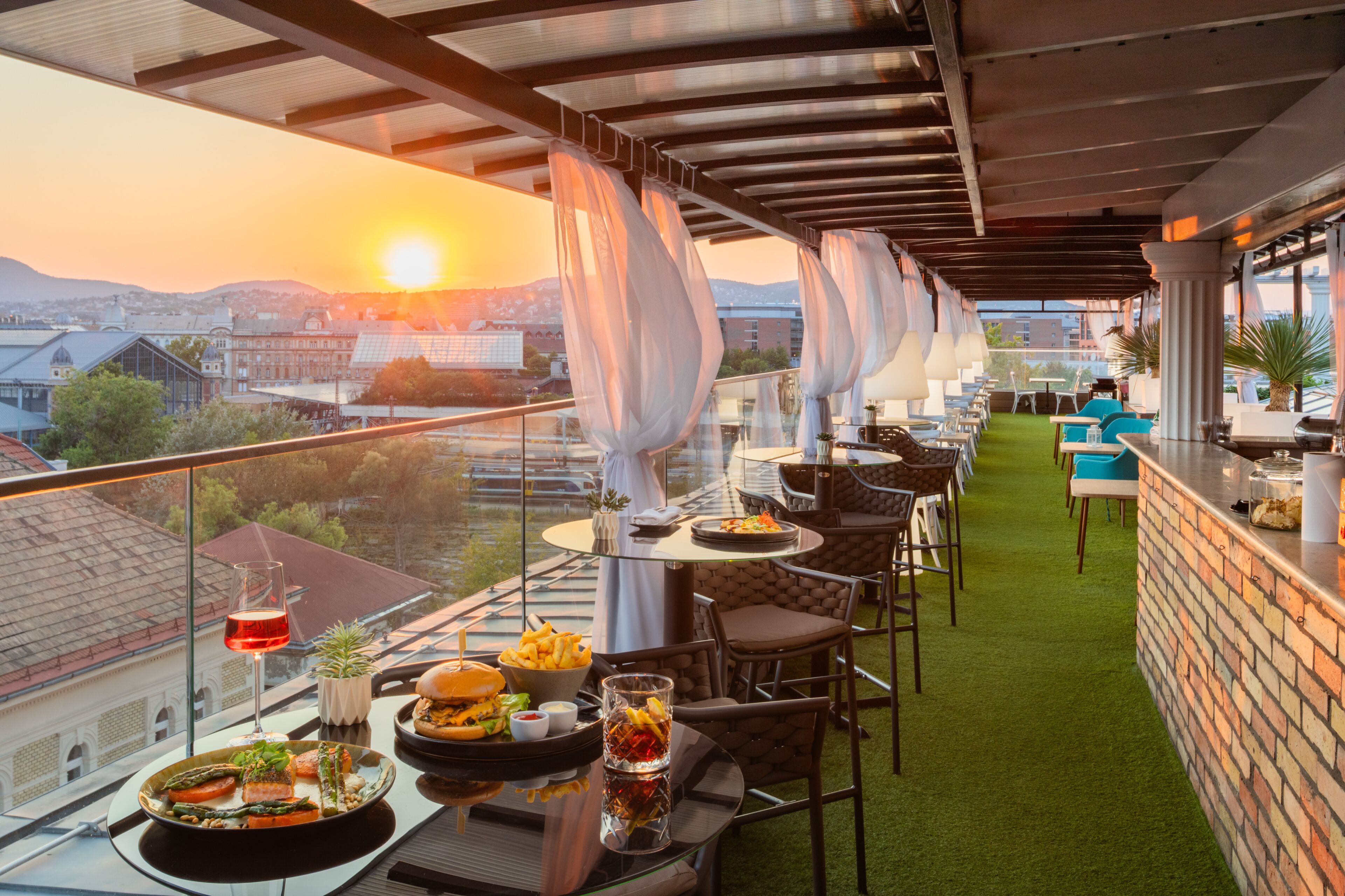 A rooftop dining area with tables and chairs, a view of the cityscape, and a sunset in the background.