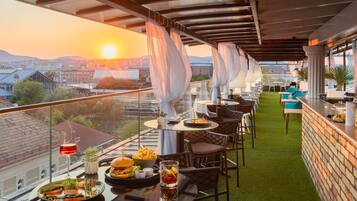 A rooftop dining area with tables and chairs, a view of the cityscape, and a sunset in the background.