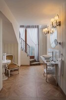 A bathroom with a large mirror, a sink, and a chair. There is a staircase leading to a balcony with curtains.