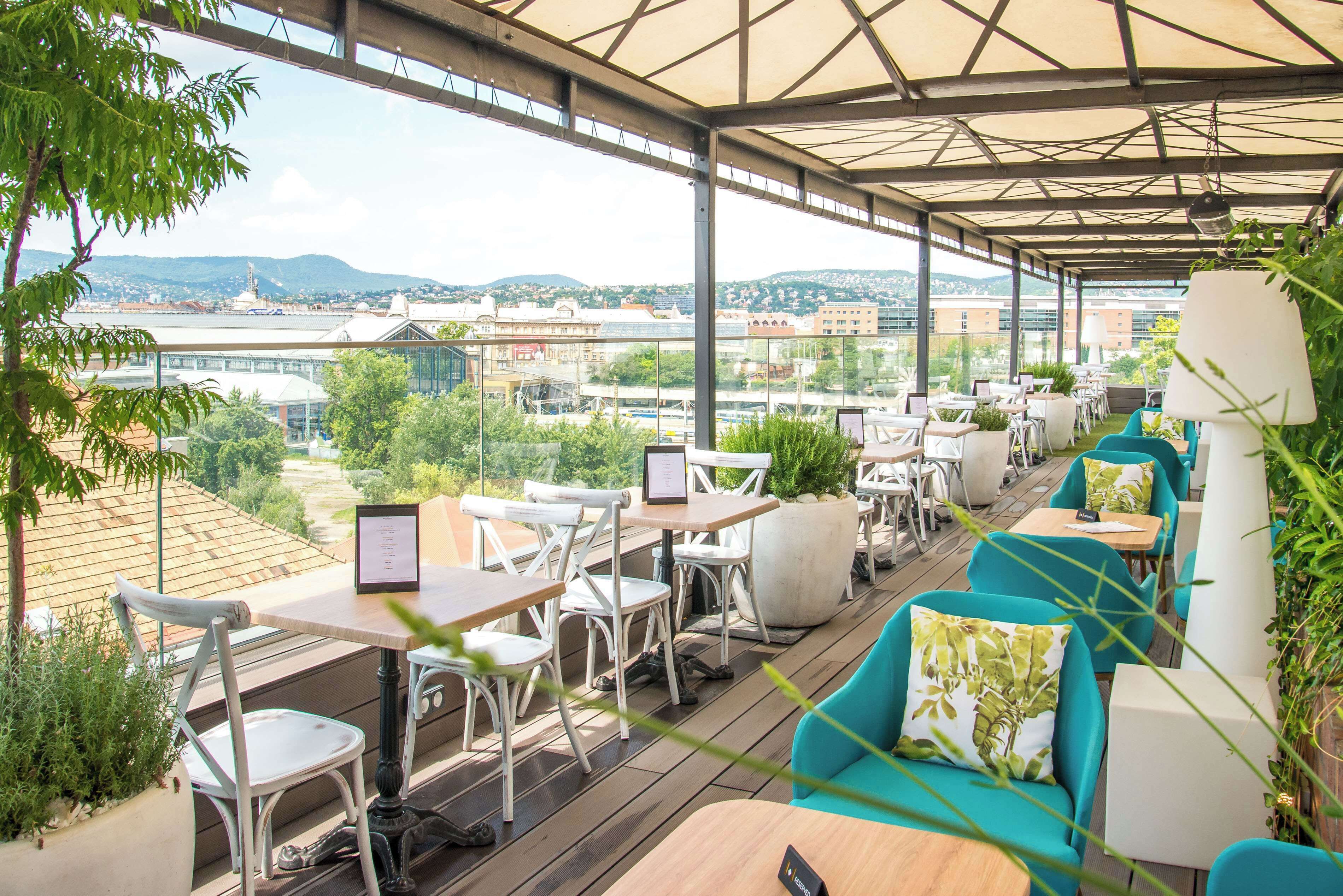 An outdoor café with tables and chairs, large windows, and potted plants.