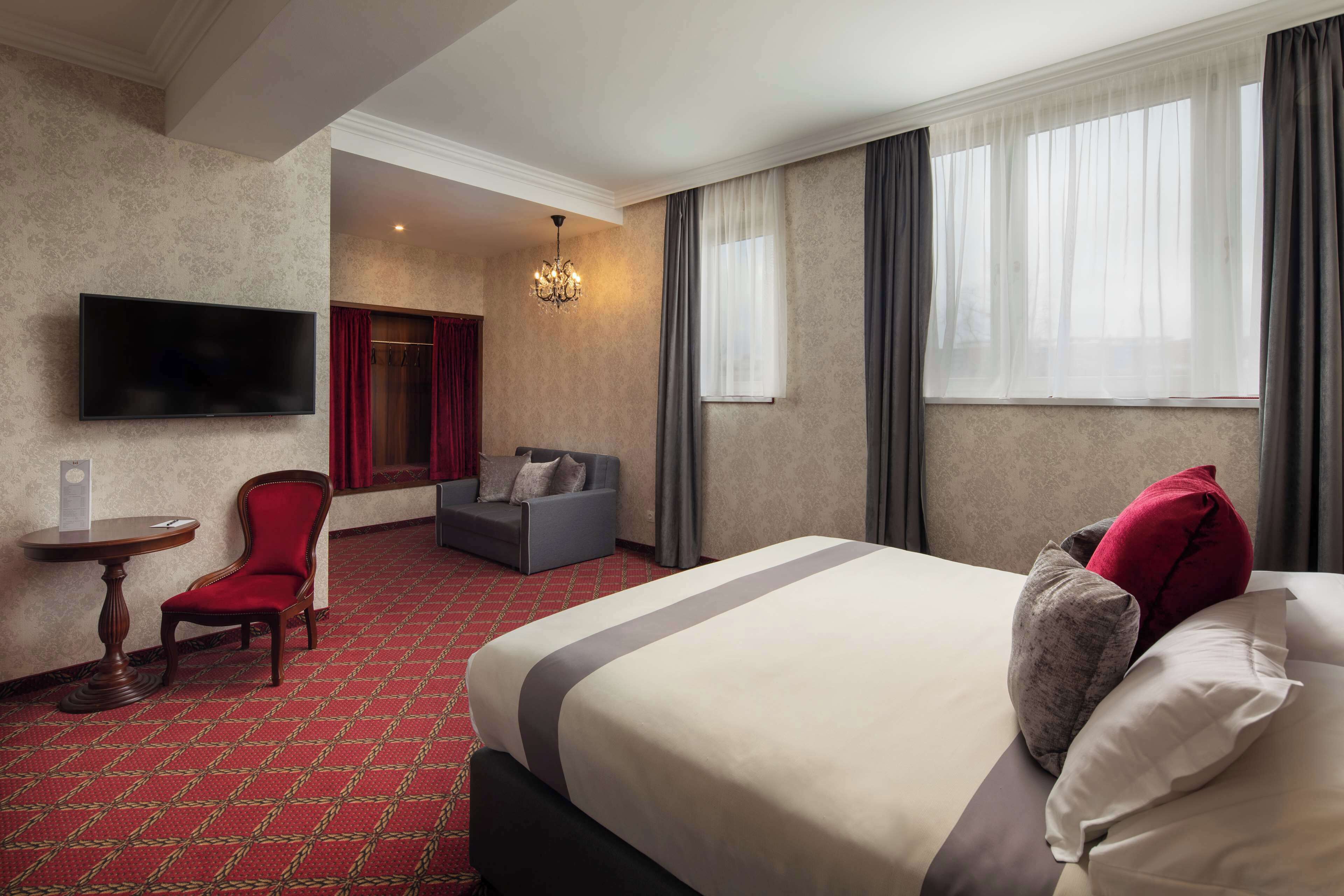 A hotel room with a large bed, a TV mounted on the wall, a small table, a red chair, a grey sofa, and a chandelier.