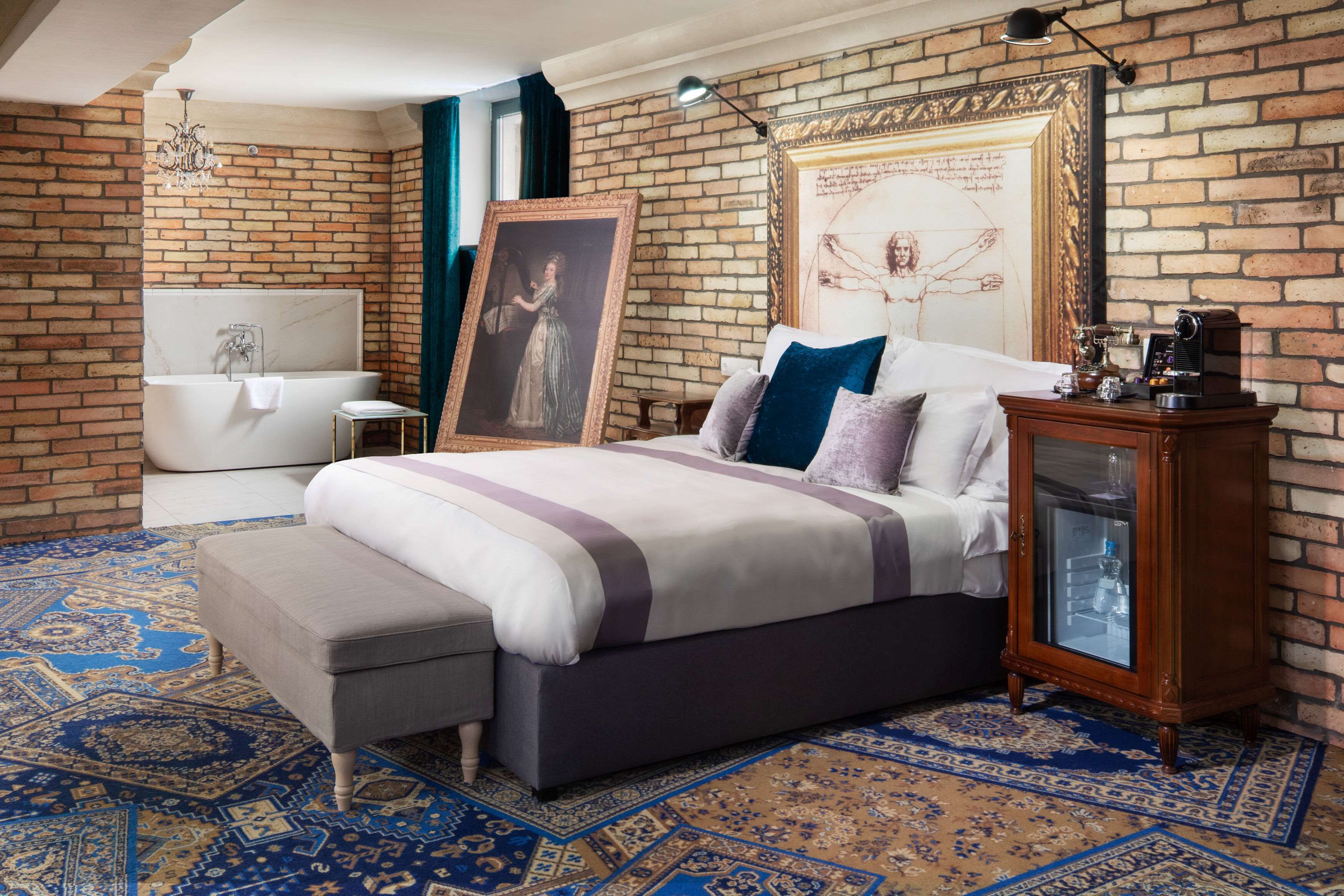 Une chambre avec un mur en briques, un grand lit, une table de chevet, une commode, un lustre et un tableau encadré accroché au mur.