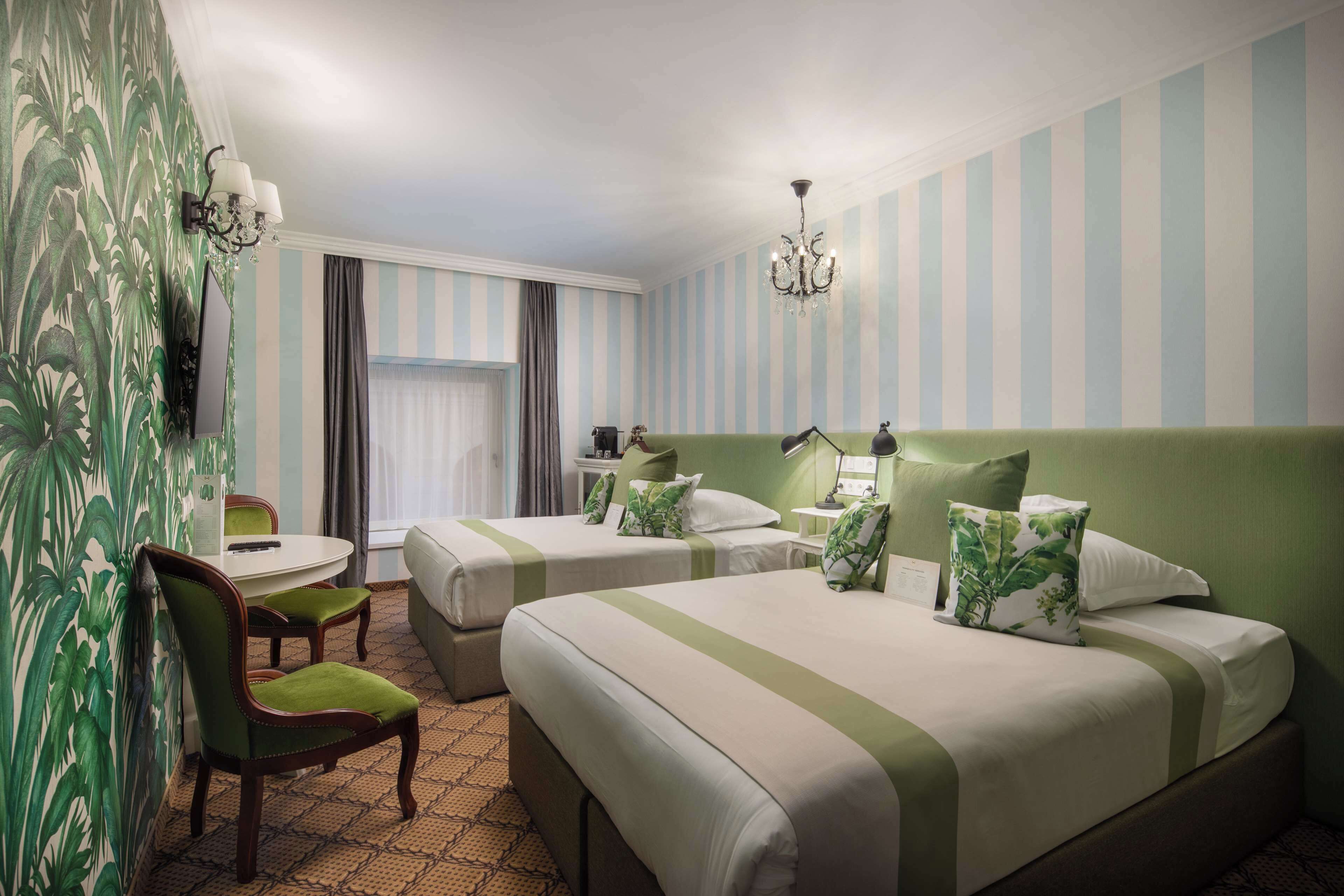 Ein Hotelzimmer mit zwei Betten, einem Schreibtisch, einem Stuhl, einer Kandelaber und gestreifter Tapete.