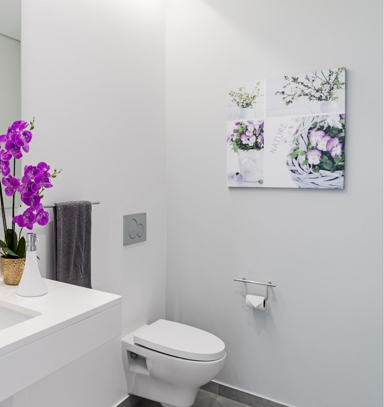 A modern bathroom with a white sink, toilet, and a wall-mounted toilet paper holder. There is a potted orchid on the counter and a framed floral artwork above the toilet.