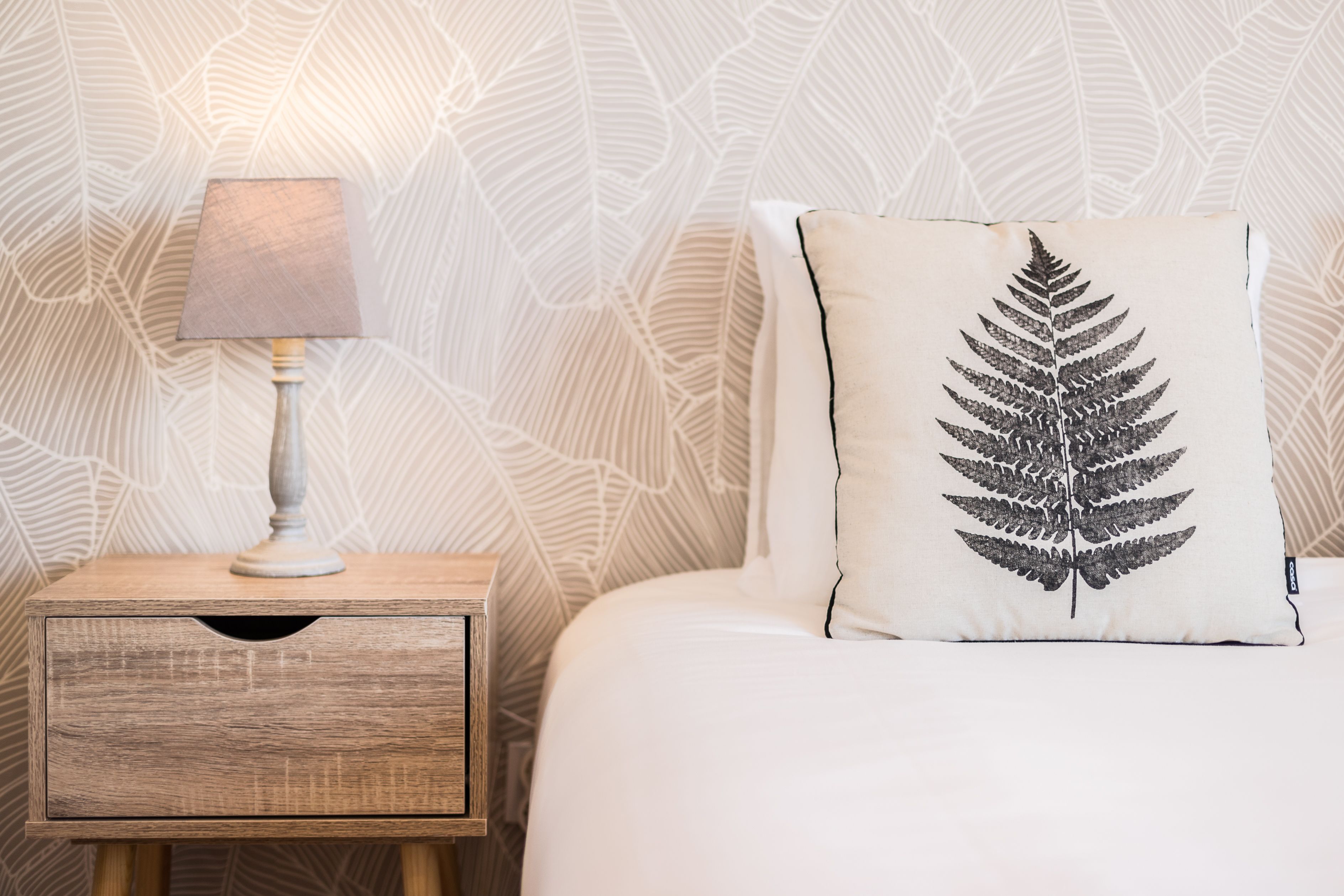 A neatly made bed with a pillow featuring a fern design, a bedside lamp on a wooden nightstand, and a patterned wallpaper.