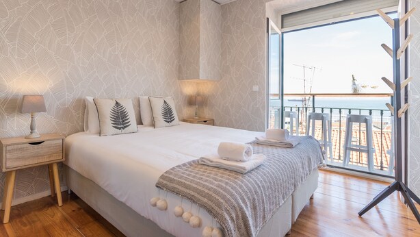 A bedroom with a large bed, a nightstand, a lamp, and a view of the beach through a balcony. - Alfama Vintage Studio Apartment w/ RiverView - by LU Holidays (Lisbon)