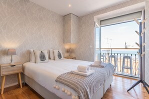 A bedroom with a large bed, a nightstand, a lamp, and a view of the beach through a balcony. - Alfama Vintage Studio Apartment w/ RiverView - by LU Holidays (Lisbon)