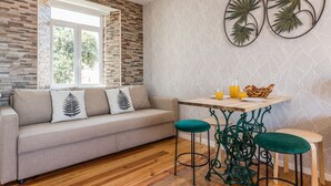 A modern living room with a stone wall, a sofa with decorative pillows, a wooden table with a basket and glass, and two green bar stools. - Alfama Vintage Studio Apartment w/ RiverView - by LU Holidays (Lisbon)