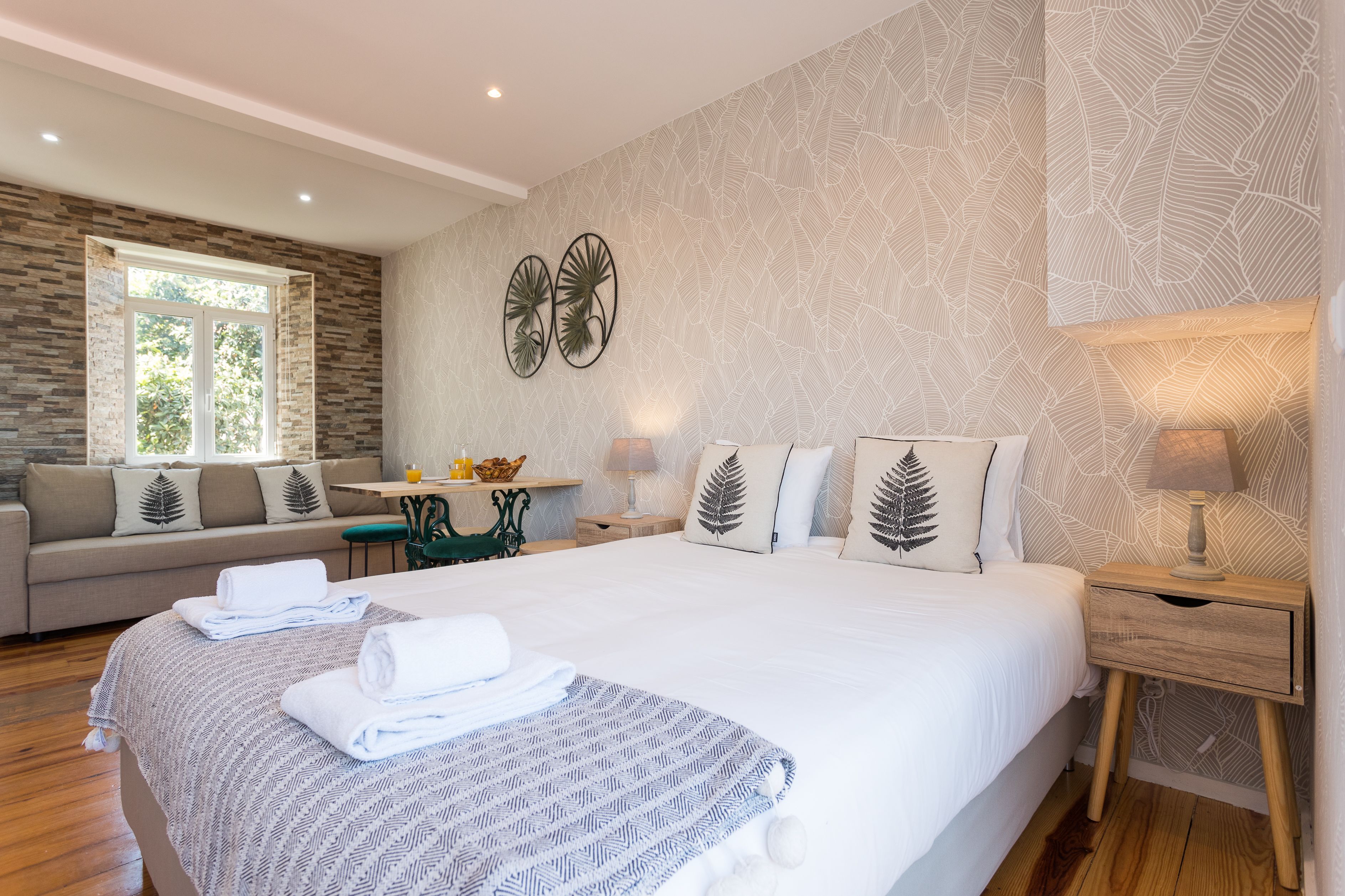 A modern hotel room with a large bed, a sofa, a small table, and a window with a view of greenery.