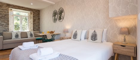 A modern hotel room with a large bed, a sofa, a small table, and a window with a view of greenery.