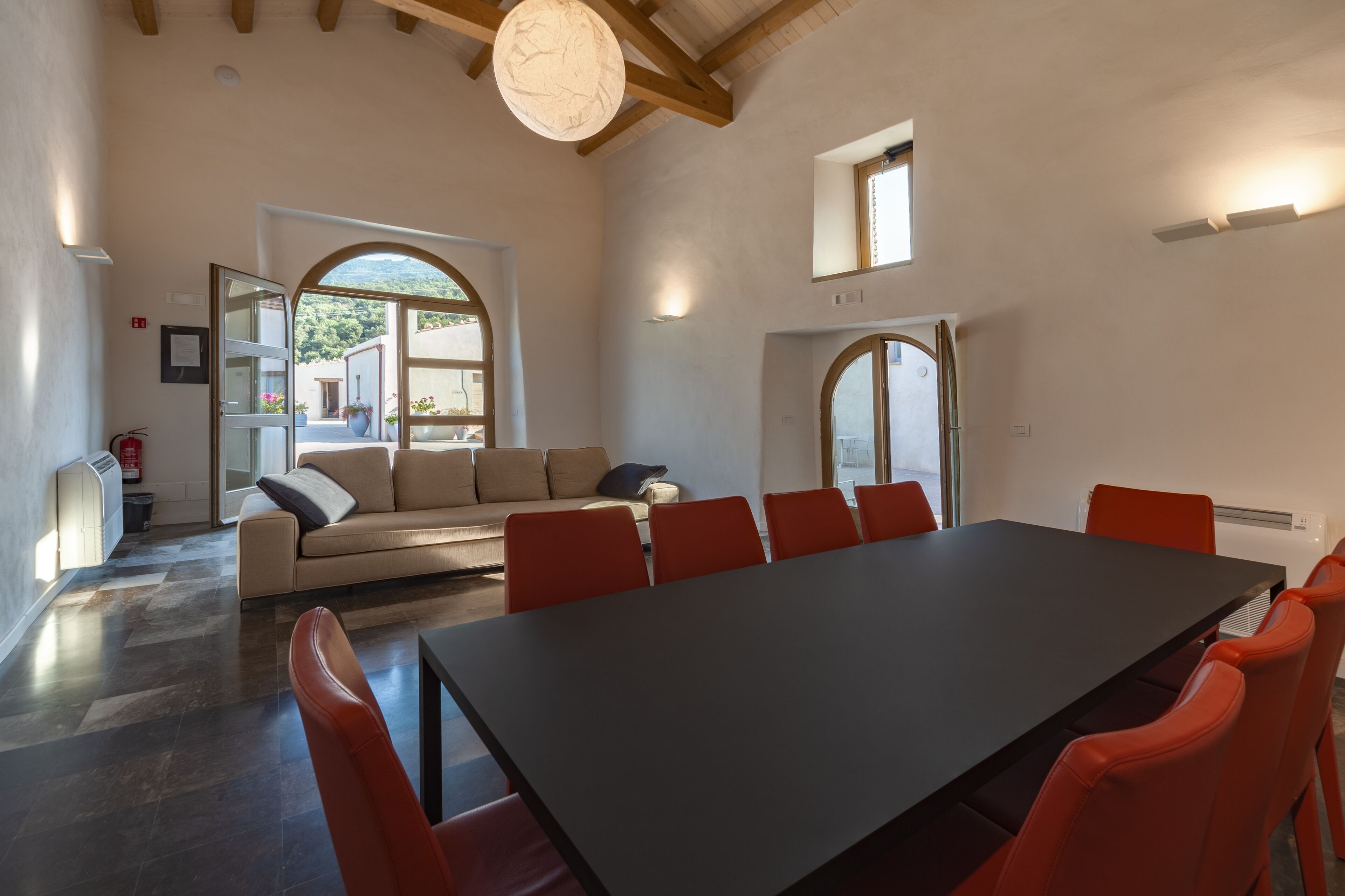 A spacious room with a large table and red chairs, a beige sofa, and a view of greenery outside.