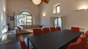 A spacious room with a large table and red chairs, a beige sofa, and a view of greenery outside.