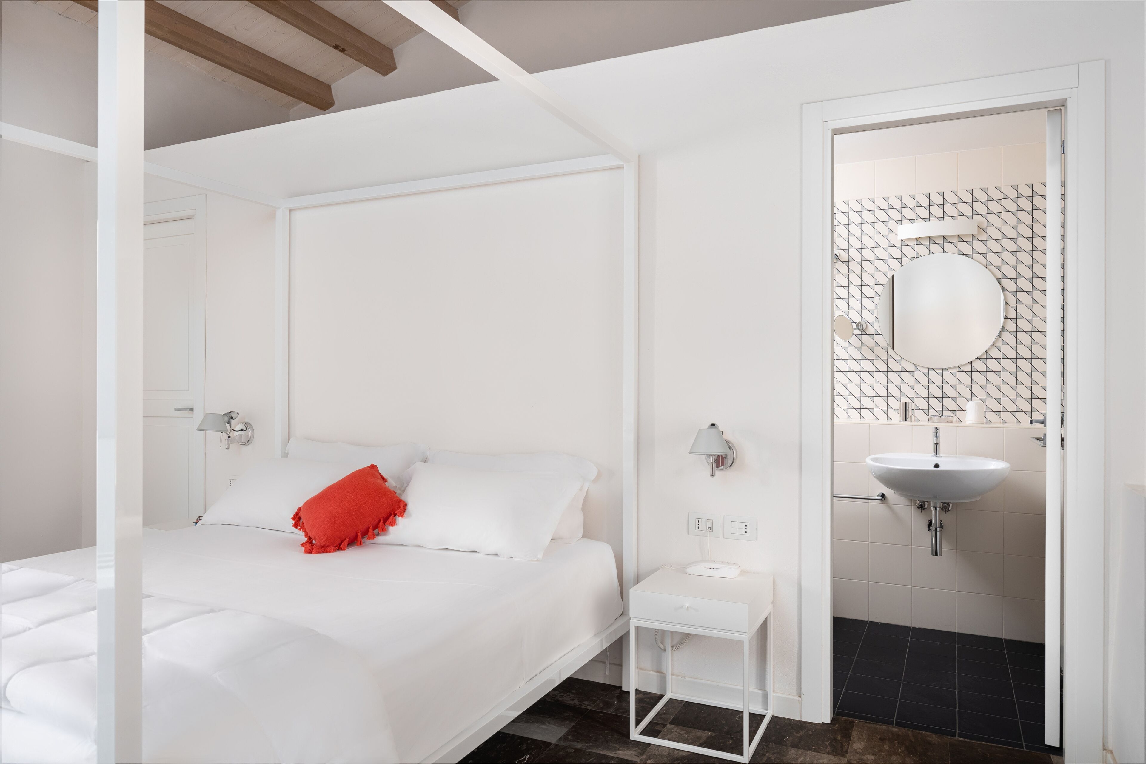 A modern hotel room with a neatly made bed, a small bedside table, a wall-mounted mirror, and a bathroom with a sink and tiled walls.