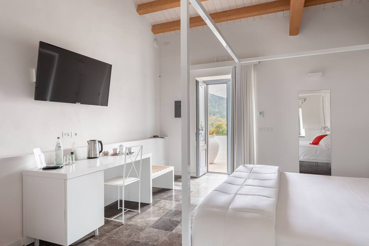 Een moderne hotelkamer met een wit bed, een kleine kitchenette, een flatscreen-televisie en een balkon met uitzicht op de bergen.