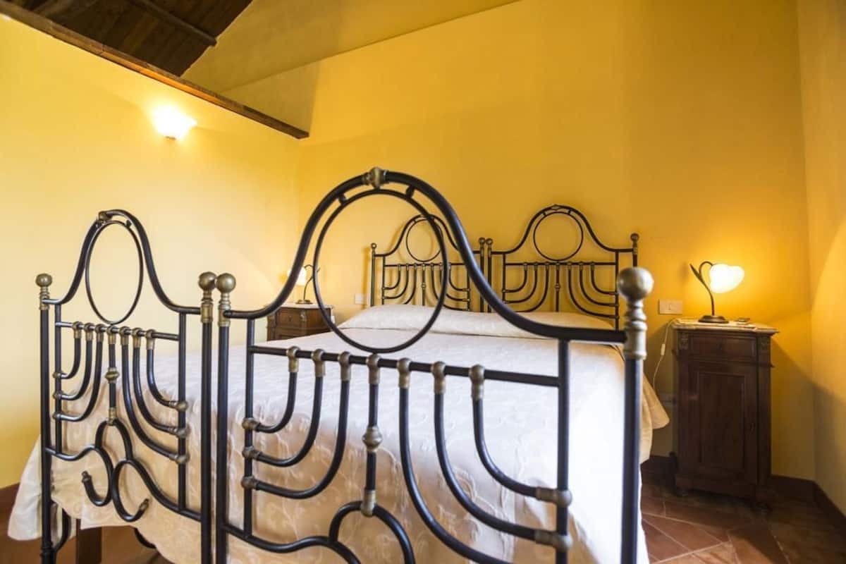 A bed with a metal frame and a headboard, a bedside table with a lamp, and a wooden cabinet against a yellow wall.