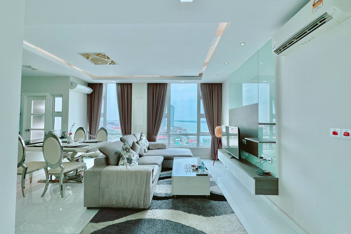 A modern living room with a sofa, coffee table, and TV. There is a dining area with chairs and a table. The room has large windows and an air conditioning unit on the wall.