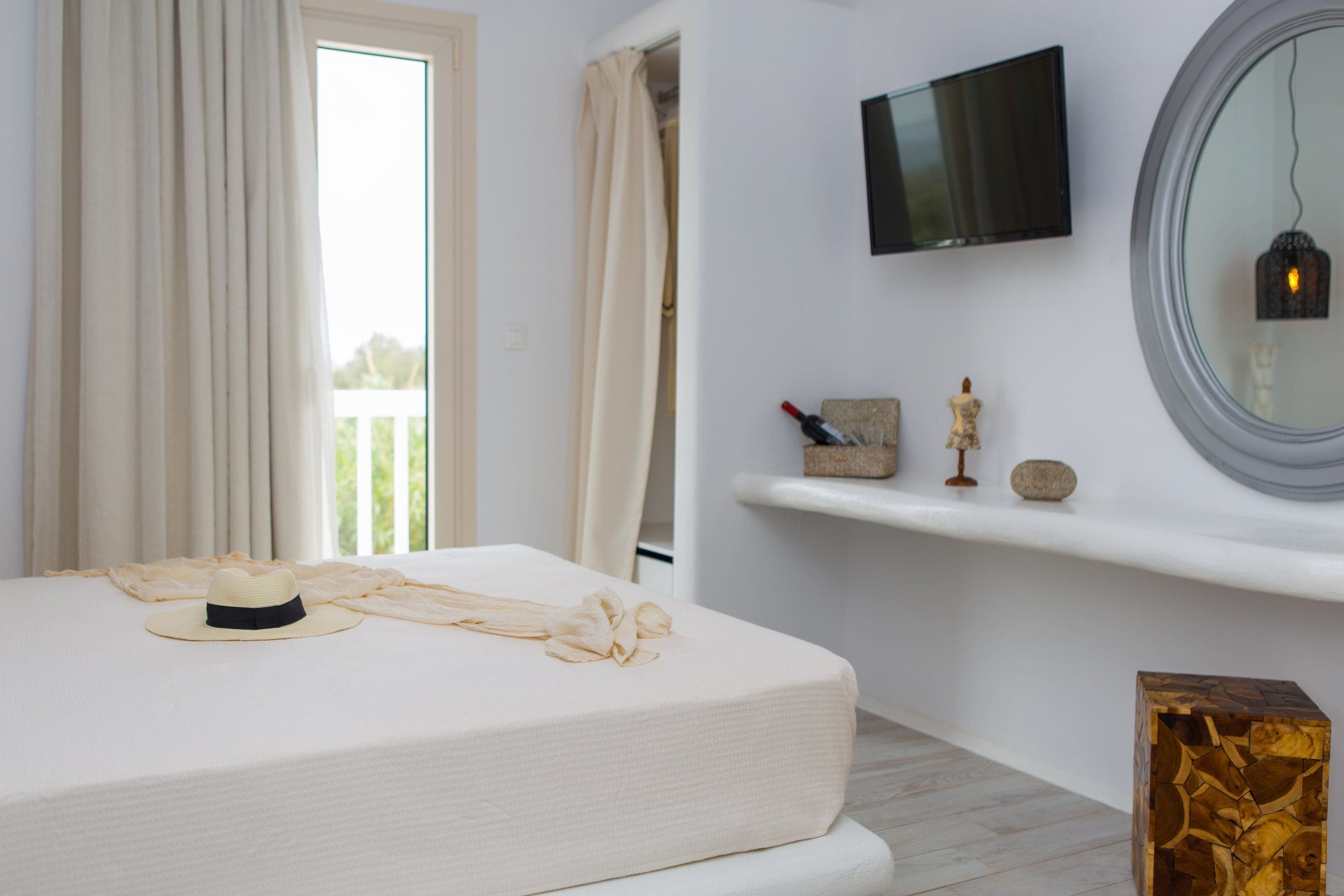 A bedroom with a bed, a TV mounted on the wall, a shelf with decorative items, and a window with curtains.