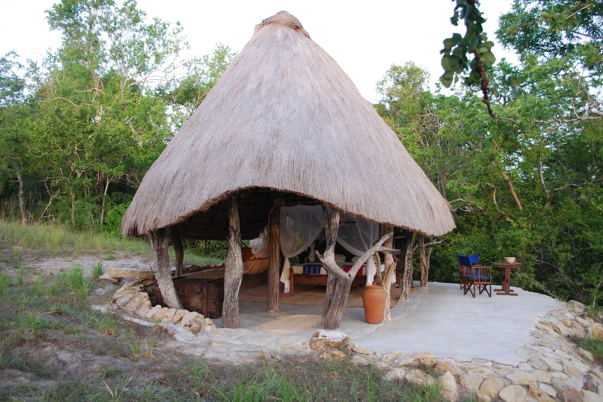 A thatched-roof hut with a bed and a table, surrounded by a stone wall and trees.