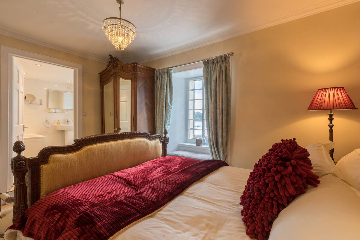 A bedroom with a large bed, a chandelier, a window with curtains, and a bedside lamp.