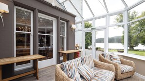 A cozy indoor seating area with wicker sofas and a wooden bench, large windows offering a view of a lake and trees. - Altskeith Country House on Loch Ard (Stirling)