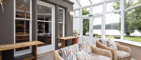 A cozy indoor seating area with wicker sofas and a wooden bench, large windows offering a view of a lake and trees.