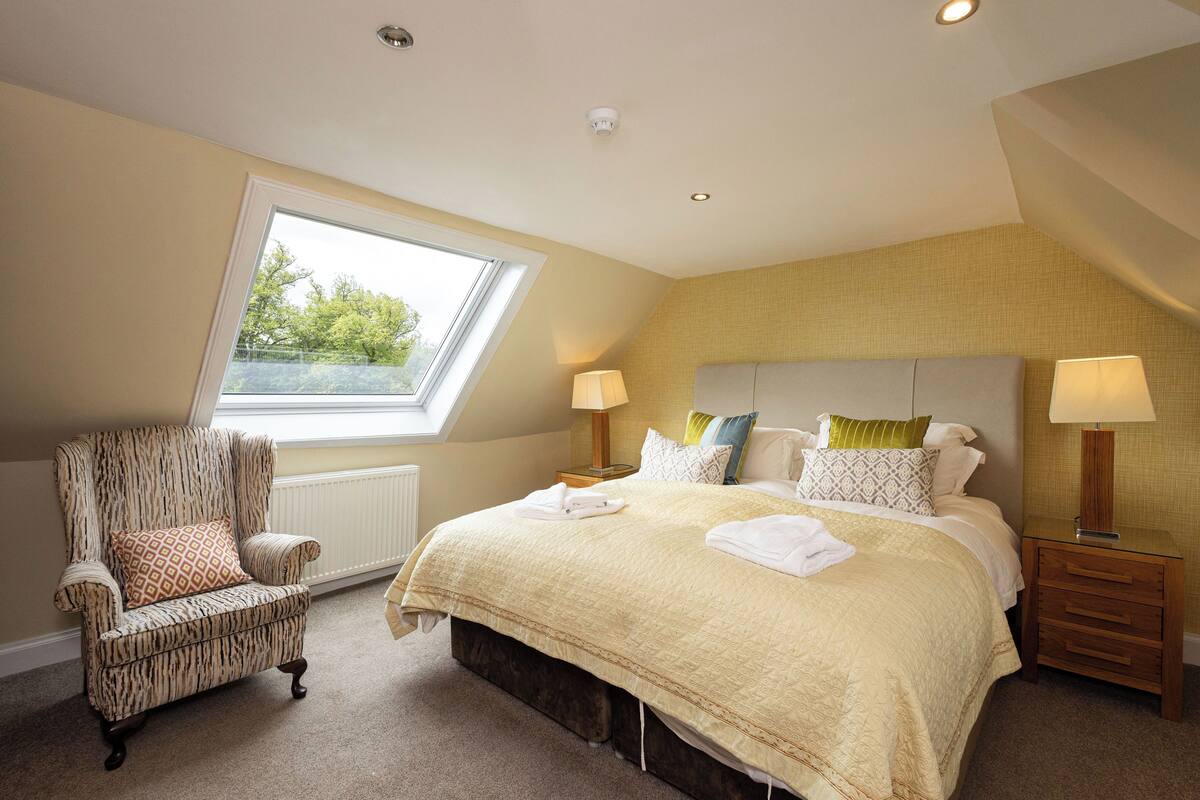 A bedroom with a large bed, a chair, two bedside tables, and a window with a view of greenery.