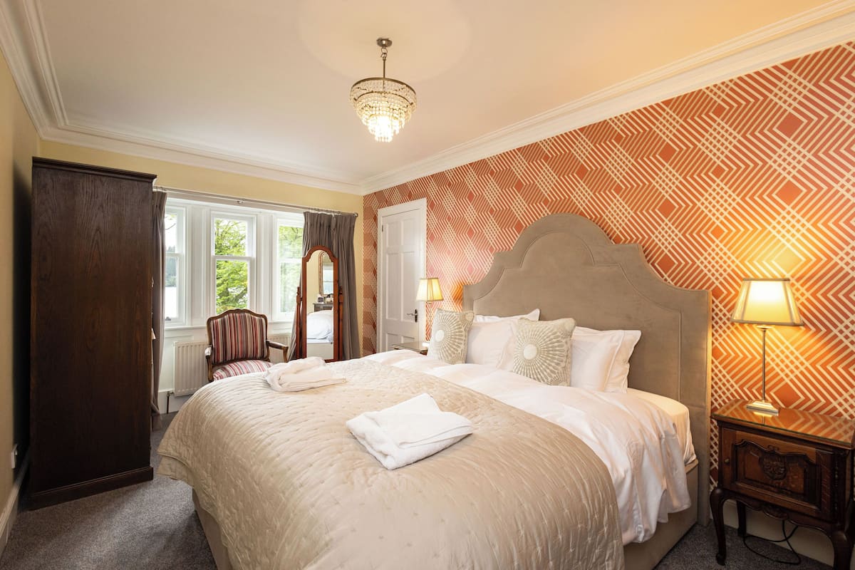 A neatly made bed with a headboard, a bedside lamp, and a window with curtains.