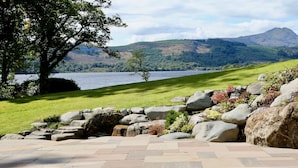 A landscaped garden with a stone wall, a grassy area, and a view of a lake and mountains. - Altskeith Country House on Loch Ard (Stirling)