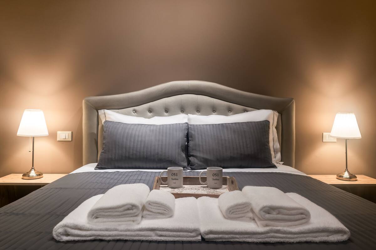 Un lit bien fait, avec une tête de lit grise, deux oreillers, un plateau avec deux tasses et deux serviettes pliées.