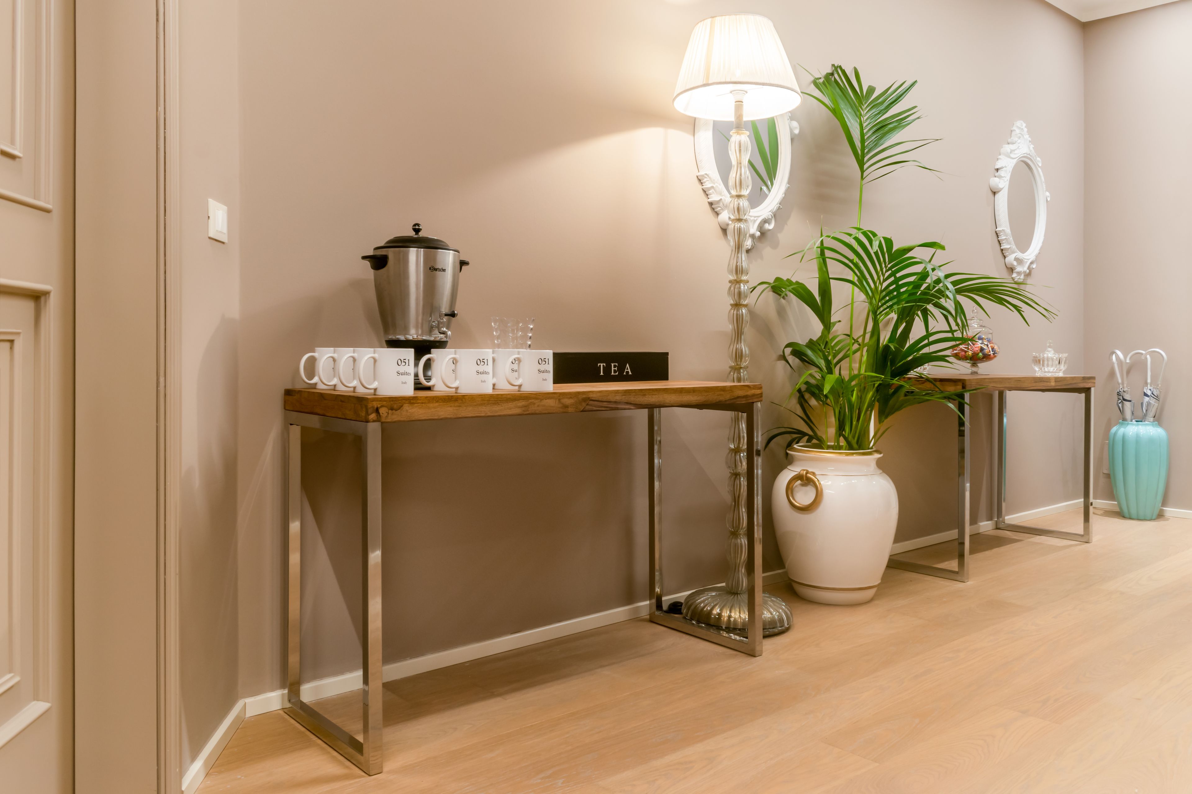 A hotel lobby with a wooden coffee table, a tea station, a potted plant, a mirror, and a suitcase.