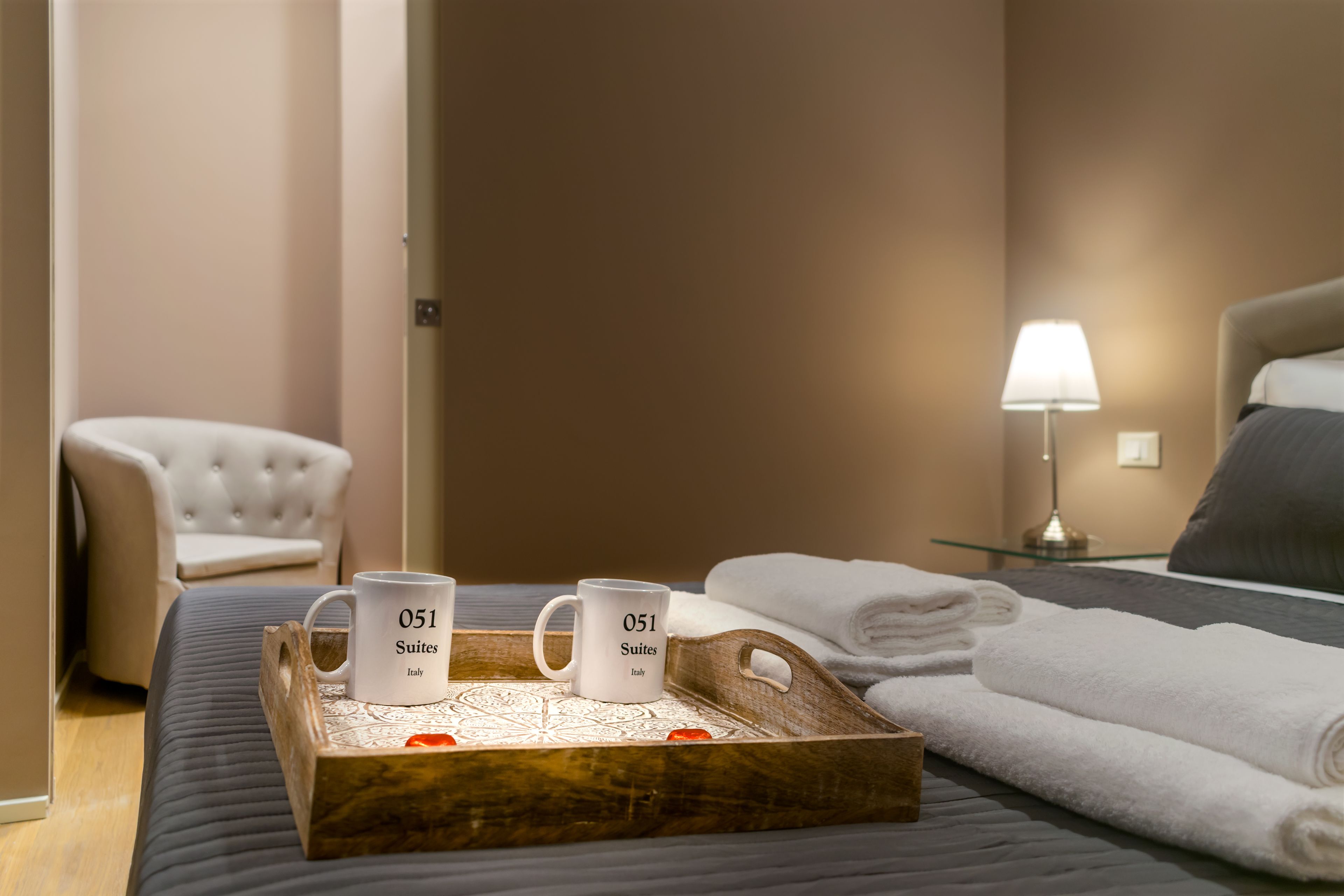 Un lit bien fait, avec des serviettes pliées, une lampe de chevet et deux tasses sur un plateau portant l’inscription « 051 Suites Italy ».