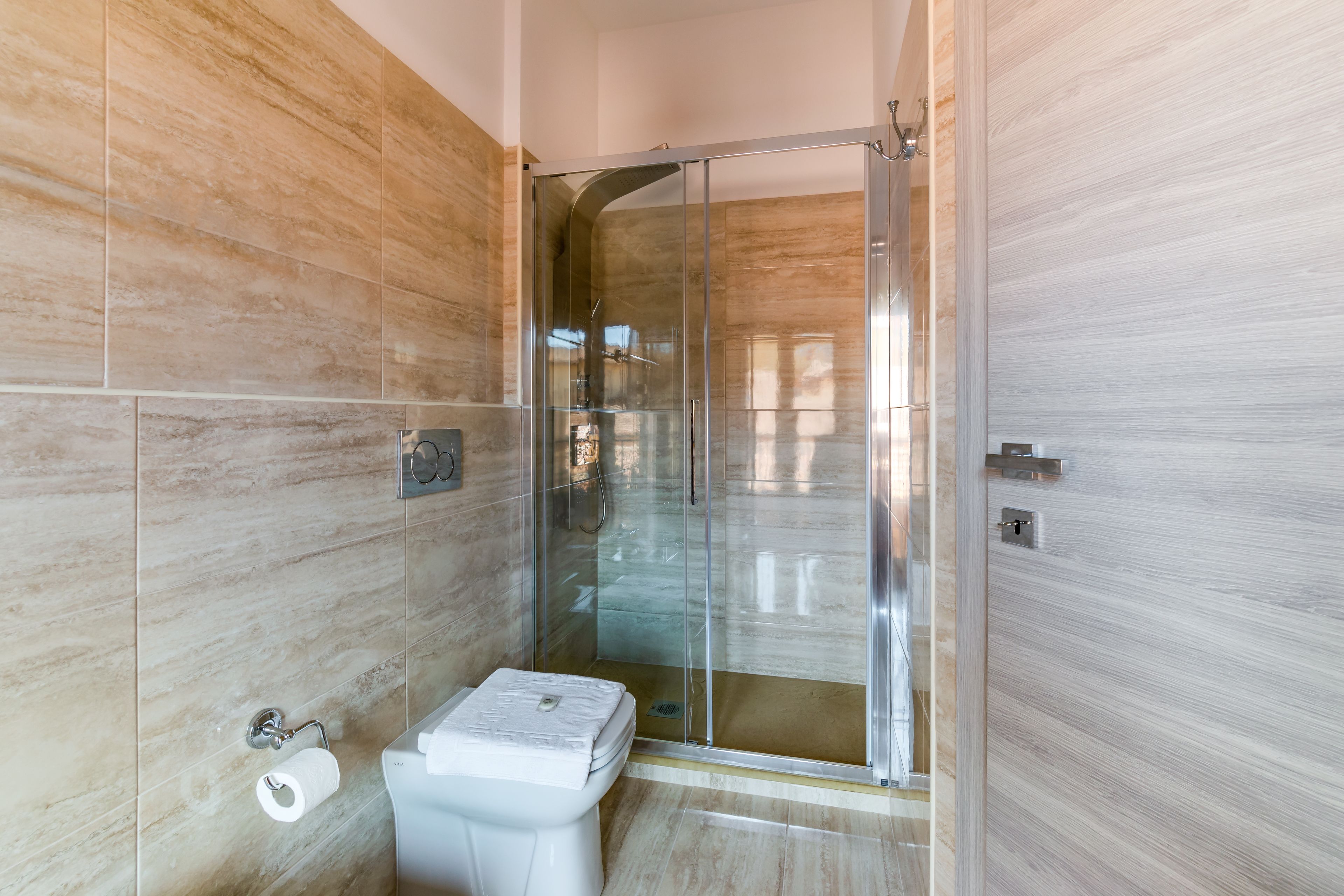 Een moderne badkamer met een douche met glazen wanden, marmeren wanden en een toilet met een papieren handdoekhouder.