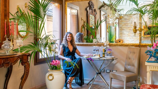 Une femme est assise à une table, devant un vase de fleurs, un miroir et une lampe décorative.