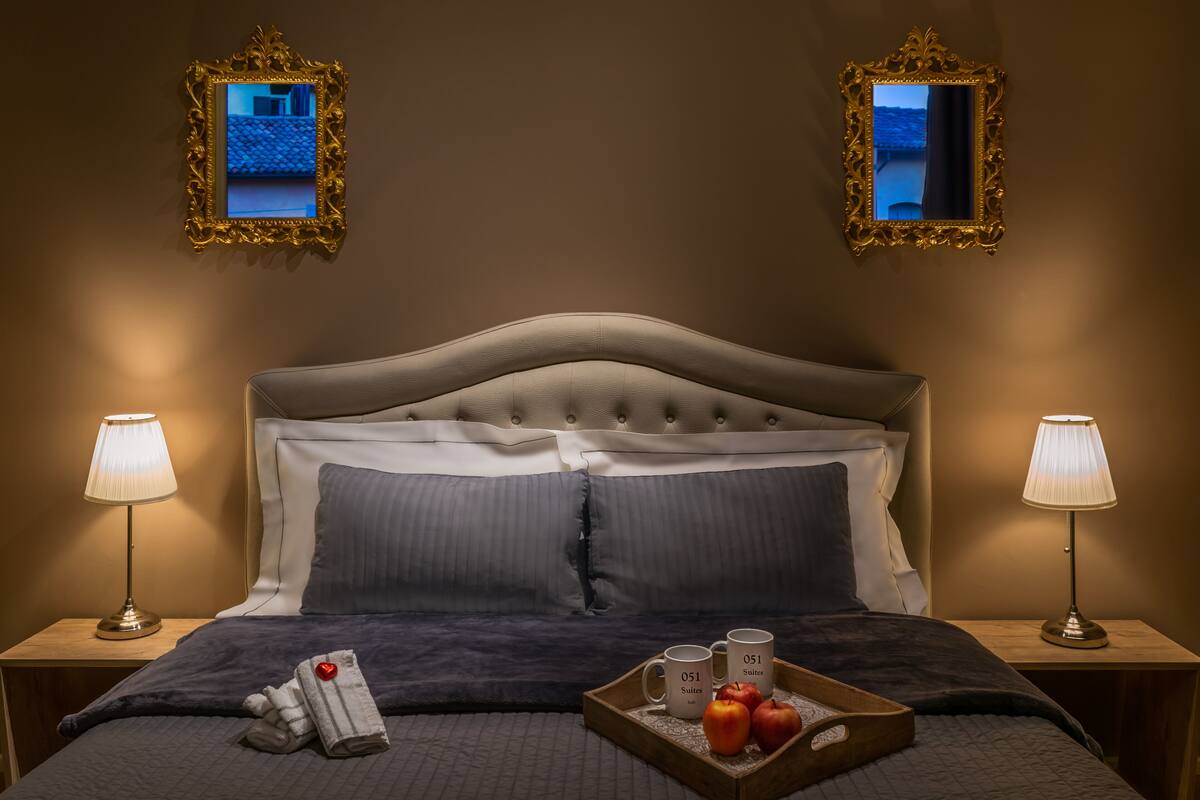 Une chambre d’hôtel avec une tête de lit capitonnée, deux lampes de chevet, un plateau avec des pommes et des tasses, et deux miroirs décoratifs accrochés aux murs.