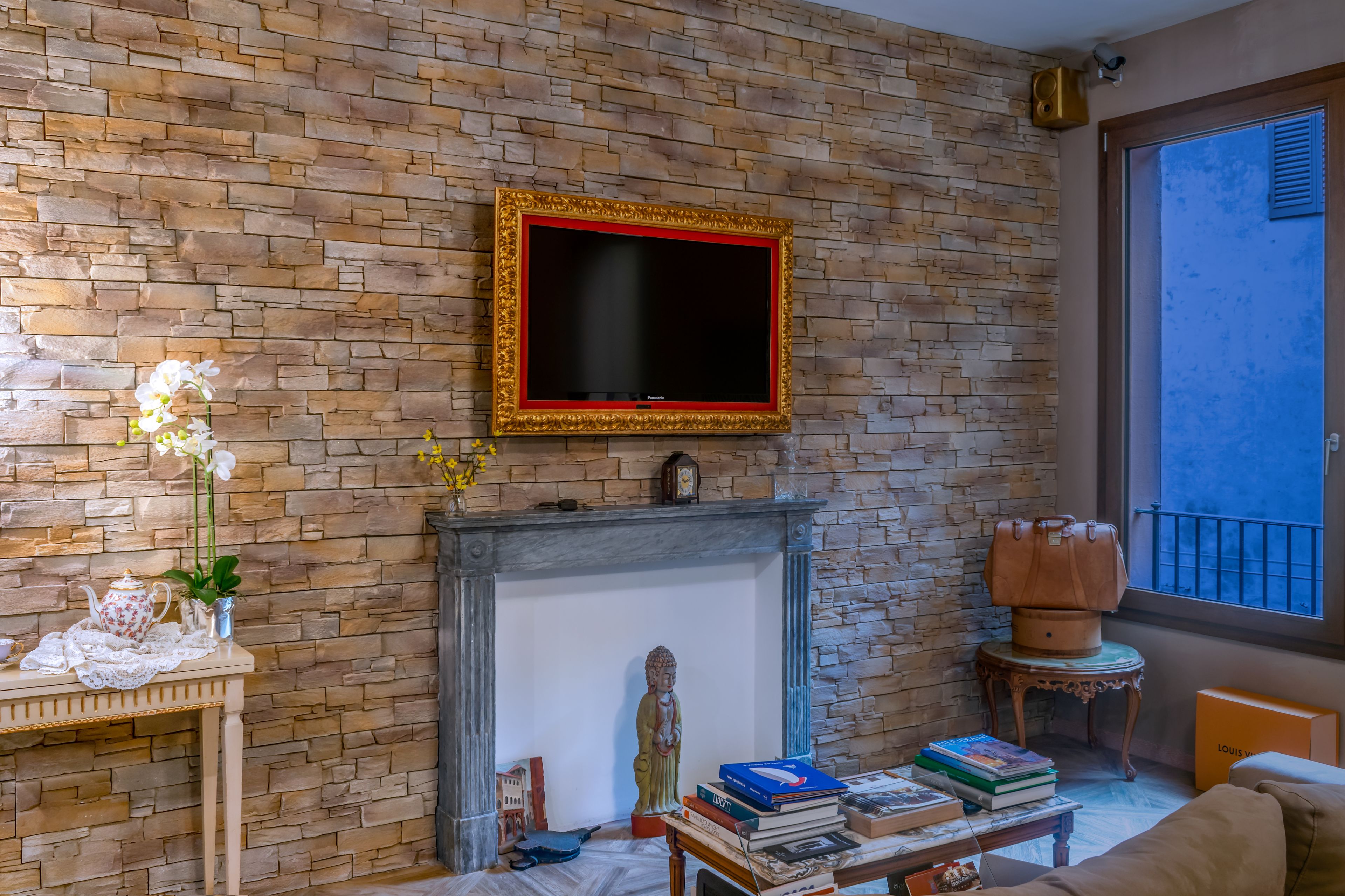 A living room with a brick wall, a fireplace, a mounted TV, a statue, a table with books, and a window with a view.