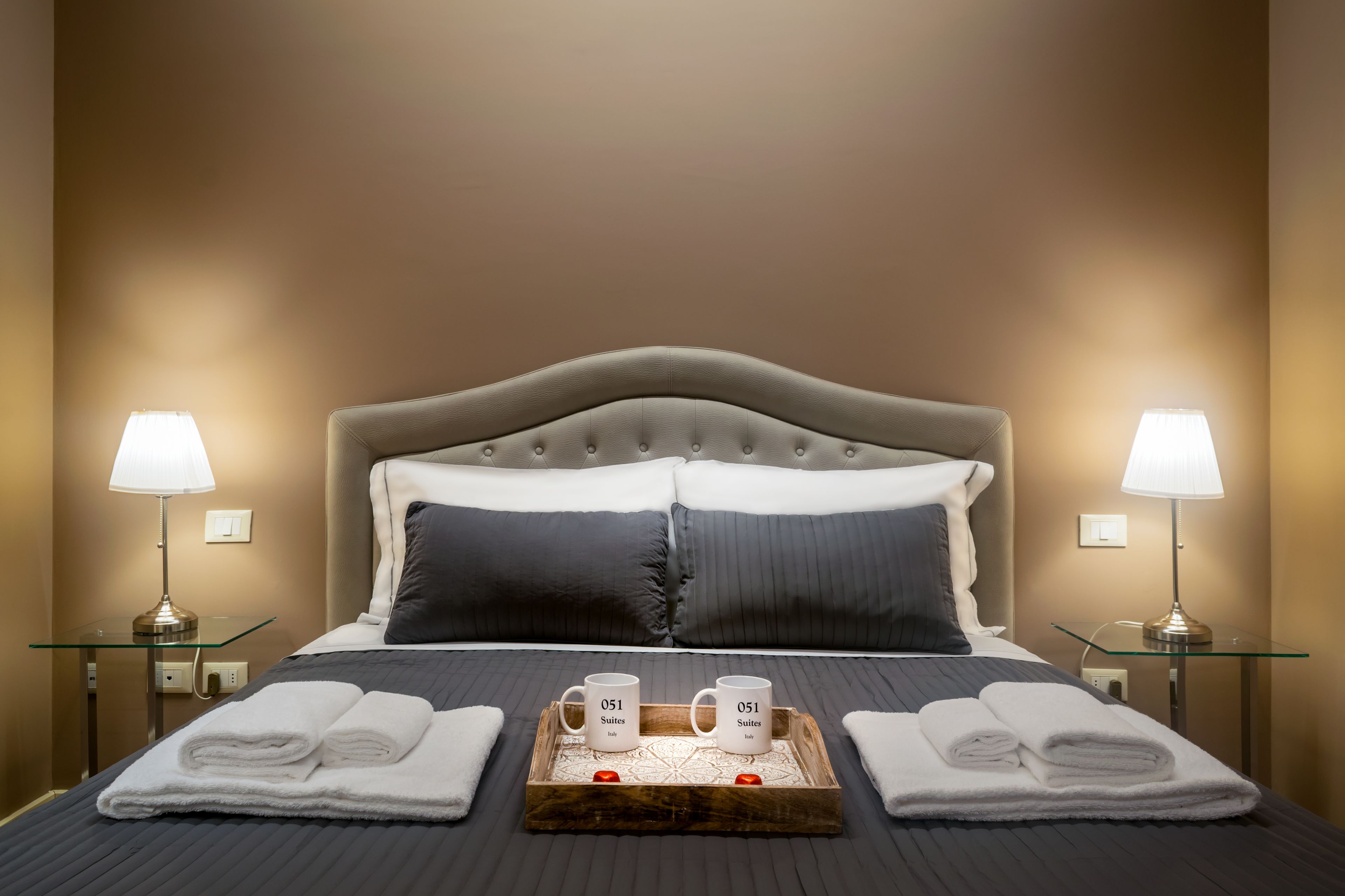 A neatly made bed with a grey headboard, two pillows, and a tray with two mugs and towels.