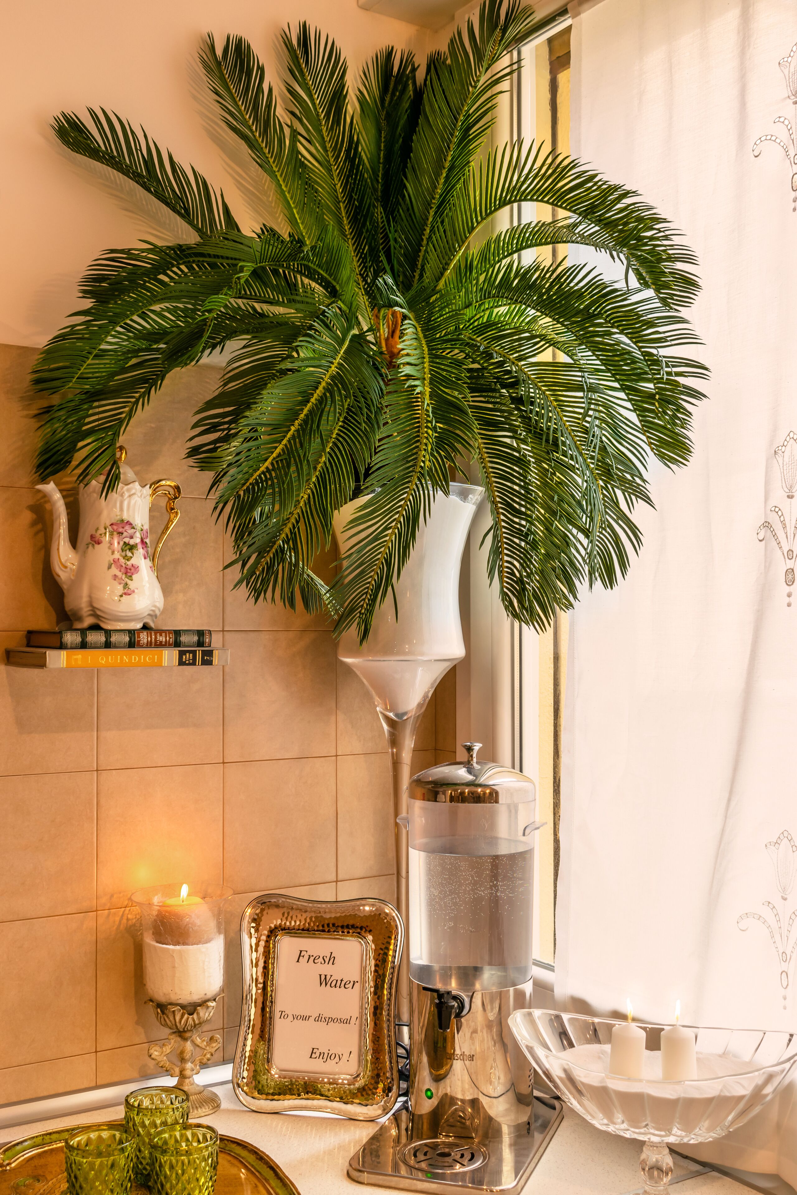 A bathroom with a potted palm plant, a white pitcher, a candle, a fresh water dispenser, and a framed sign.