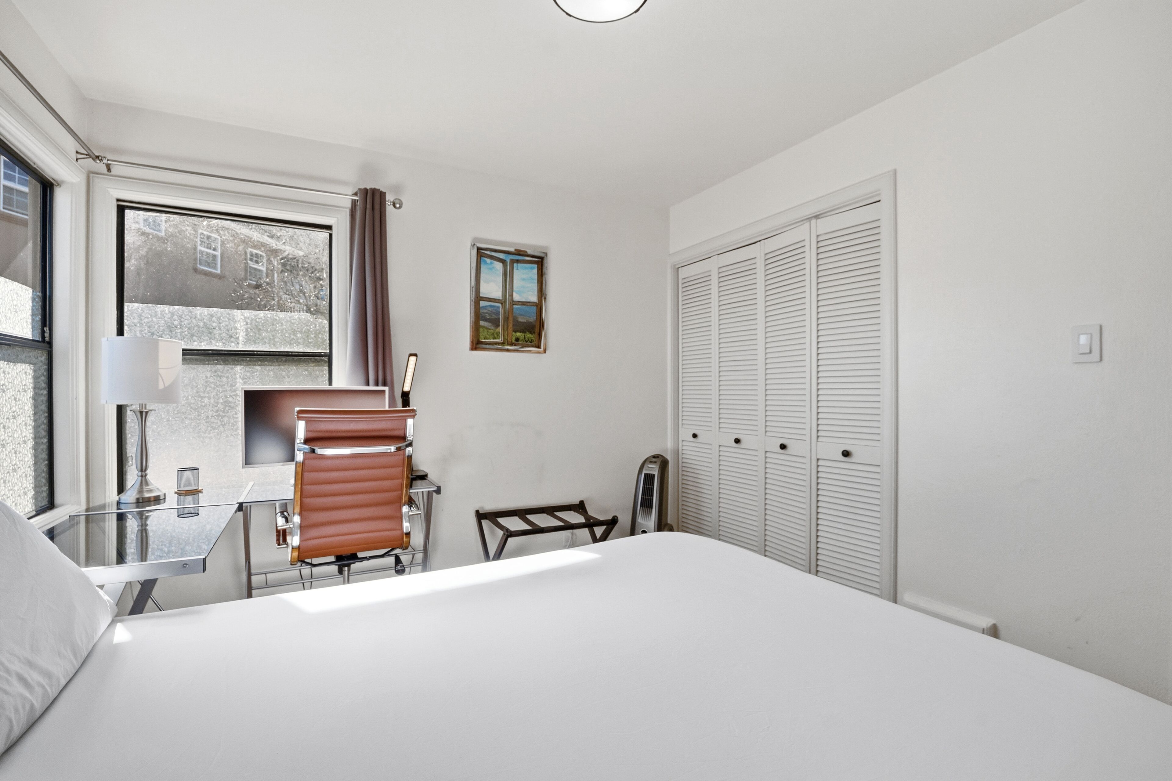 Una habitación de hotel con una cama, un escritorio con lámpara, una silla, un radiador, una ventana con cortinas y varios armarios blancos.